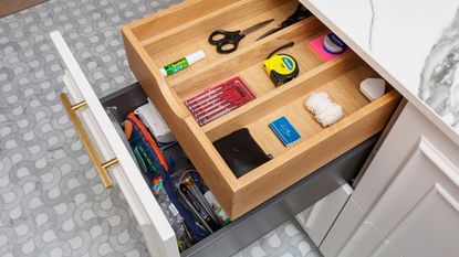 open junk drawer with scissors, screwdrivers, glue, tape, phone charger, marble countertop, white cabinetry, gray floor tiles