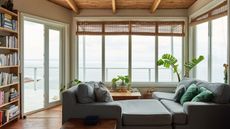 Learning how to clean windows without streaks is easy. Here is a living room with a wooden ceiling, a gray couch, and a large window with woven blinds looking onto a cloudy sky and a clear ocean