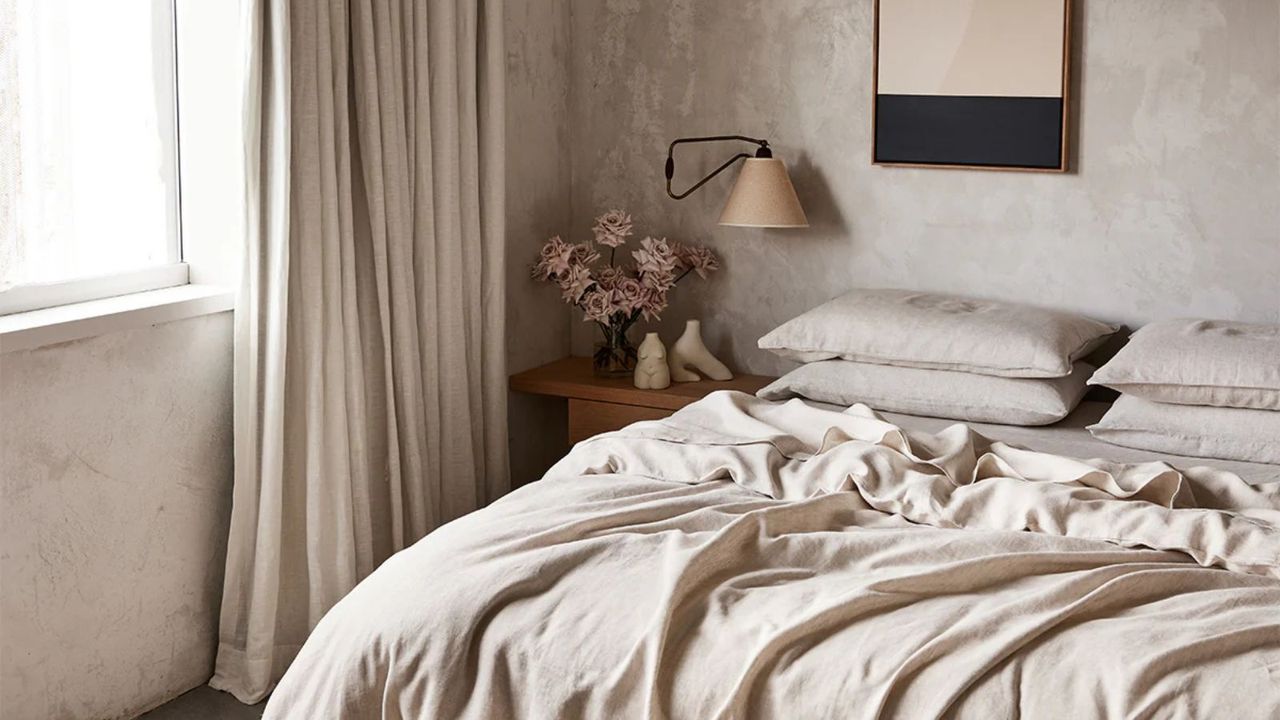 Oatmeal sheets and pillowcases on a bed beside a wooden nightstand, topped with pink floors, against beige walls. 