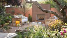 A modern garden with a bed of flowers in the foreground and an outdoor fireplace in the background and a set of garden furniture