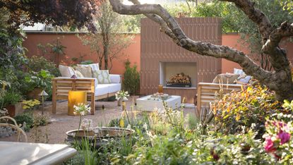 A modern garden with a bed of flowers in the foreground and an outdoor fireplace in the background and a set of garden furniture