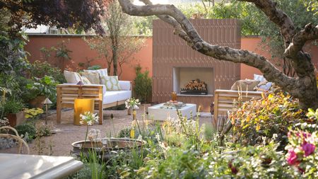 A modern garden with a bed of flowers in the foreground and an outdoor fireplace in the background and a set of garden furniture