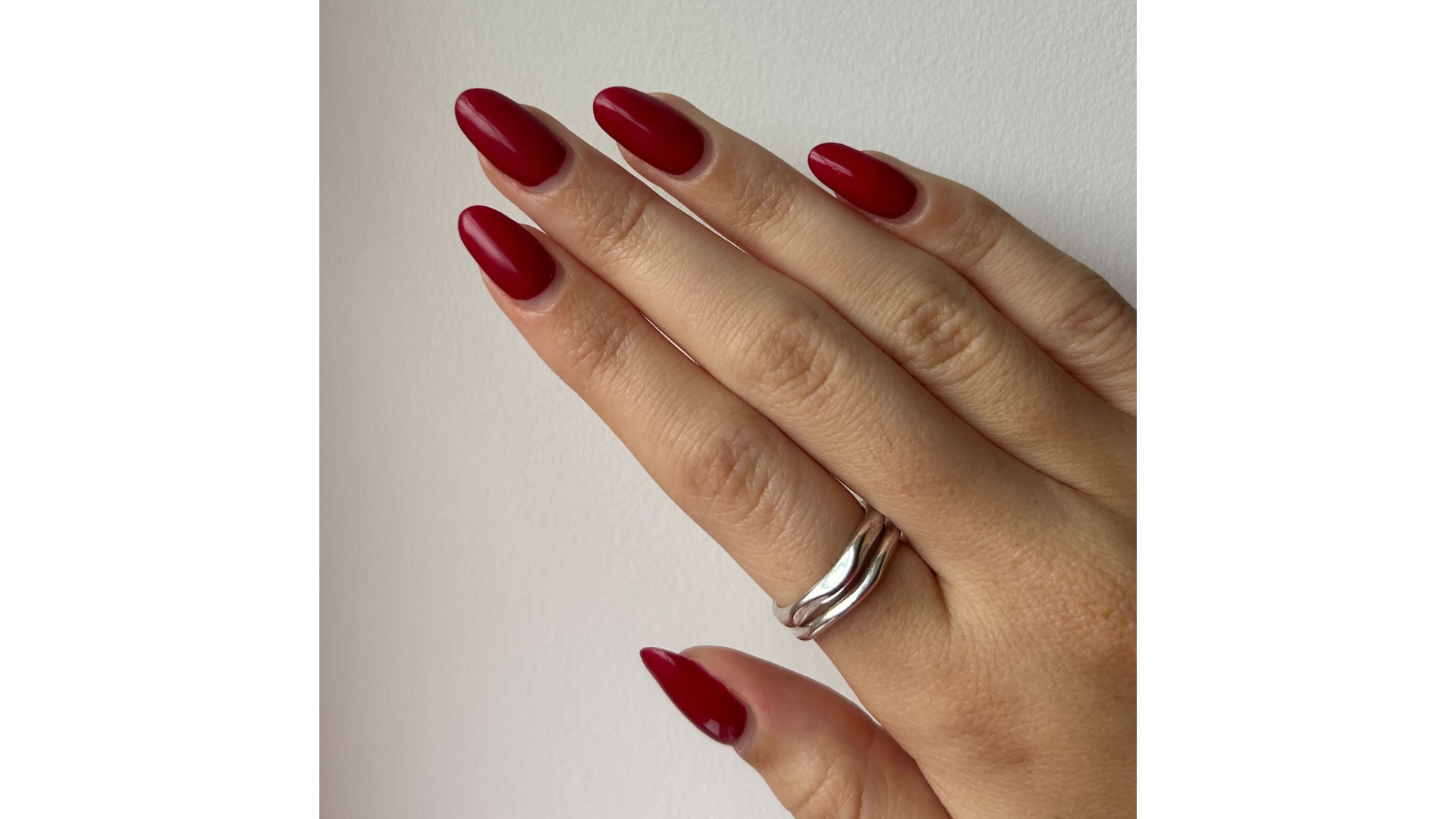 A close-up of Digital Beauty Writer Sennen Prickett's hand, pictured with long and red almond nails, in front of a white wall background