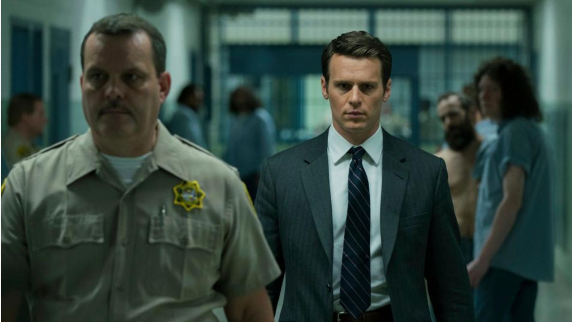 an image still from Mindhunter which shows Johnathan Groff playing Torv - an FBI agent, wearing a suit and tie being led through a prison on his way to interview a psychopath. He is being led by a police warden 