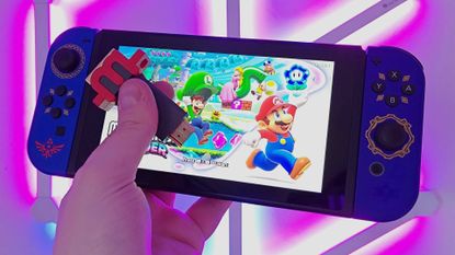 Hand holding mClassic upscaler and Nintendo Switch in one hand with Super Mario Odyssey title on screen and purple lights in backdrop.