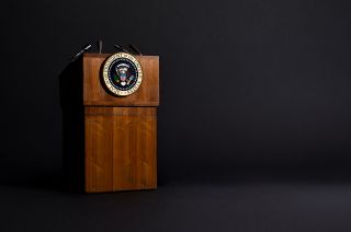 a wooden lectern with the presidential seal stands in front of a black background.