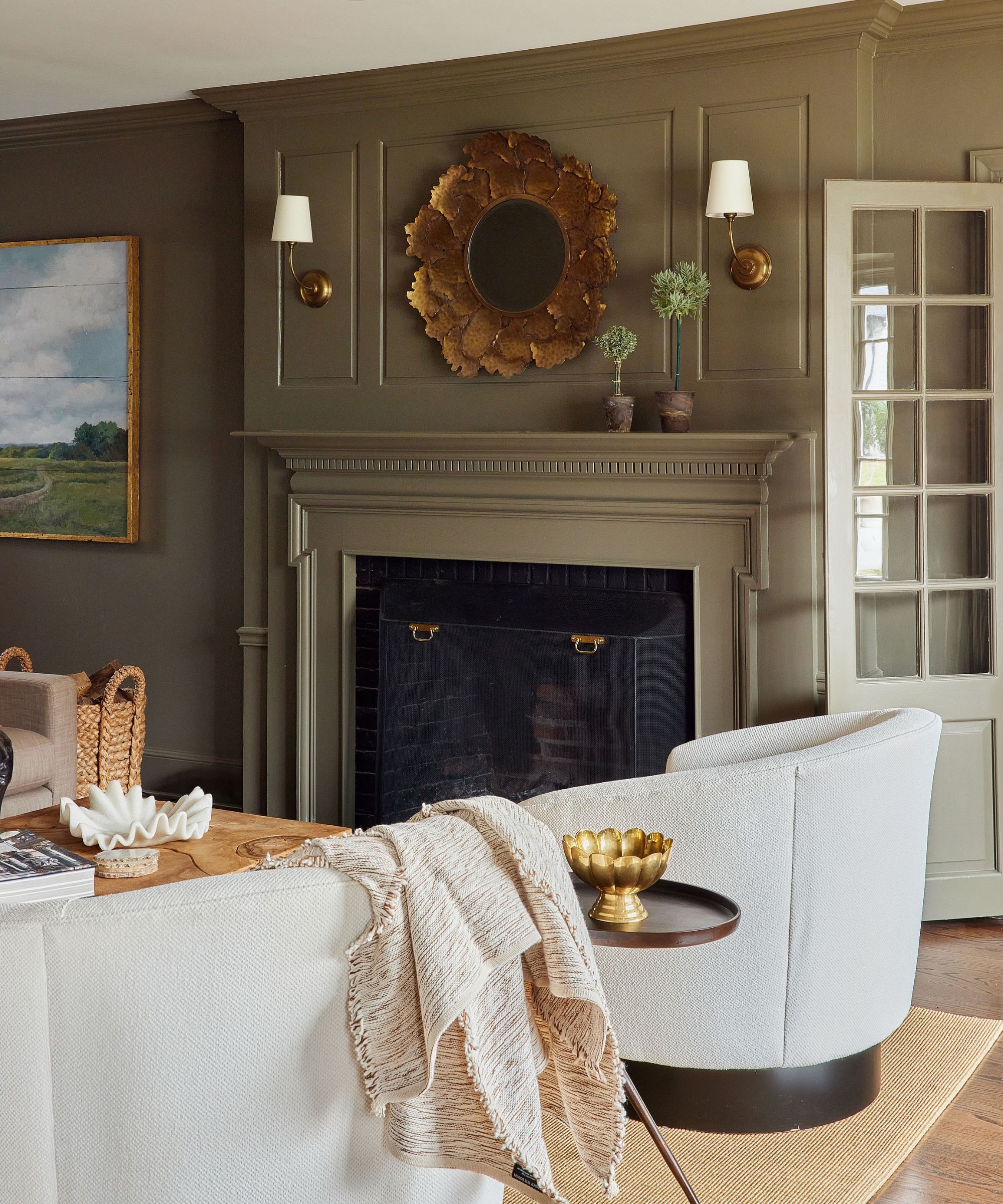 A moody living room with muddy green walls and fireplace and cream armchairs.