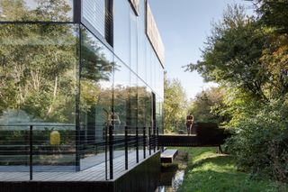 Side view of the all-glass villa. We see a deck that surrounds the villa, and the wooden bridge that leads to the entryway.