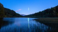 A lone meteor streaks Earthward at night, while reflected in a placid lake lined by trees.