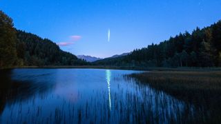 A lone meteor streaks Earthward at night, while reflected in a placid lake lined by trees.