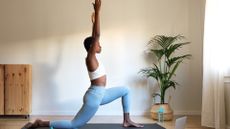 woman performing a low lunge with arms extended overhead, wearing blue leggings and white croptop in a living room setting with a plant to one side