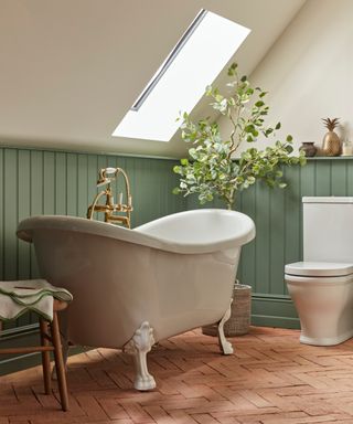 white roll top bath under eaves with terracotta brick pavier flooring and green wooden tongue and groove panelling around lower half of bathrooom