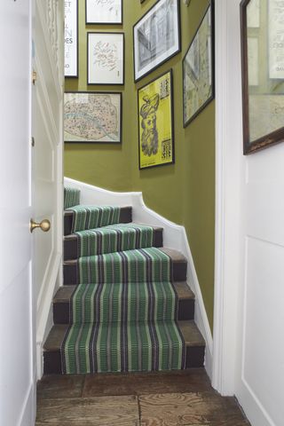 A staircase with olive green paint on the walls, black-framed wall art and green and black striped runner up stairs by Roger Oates