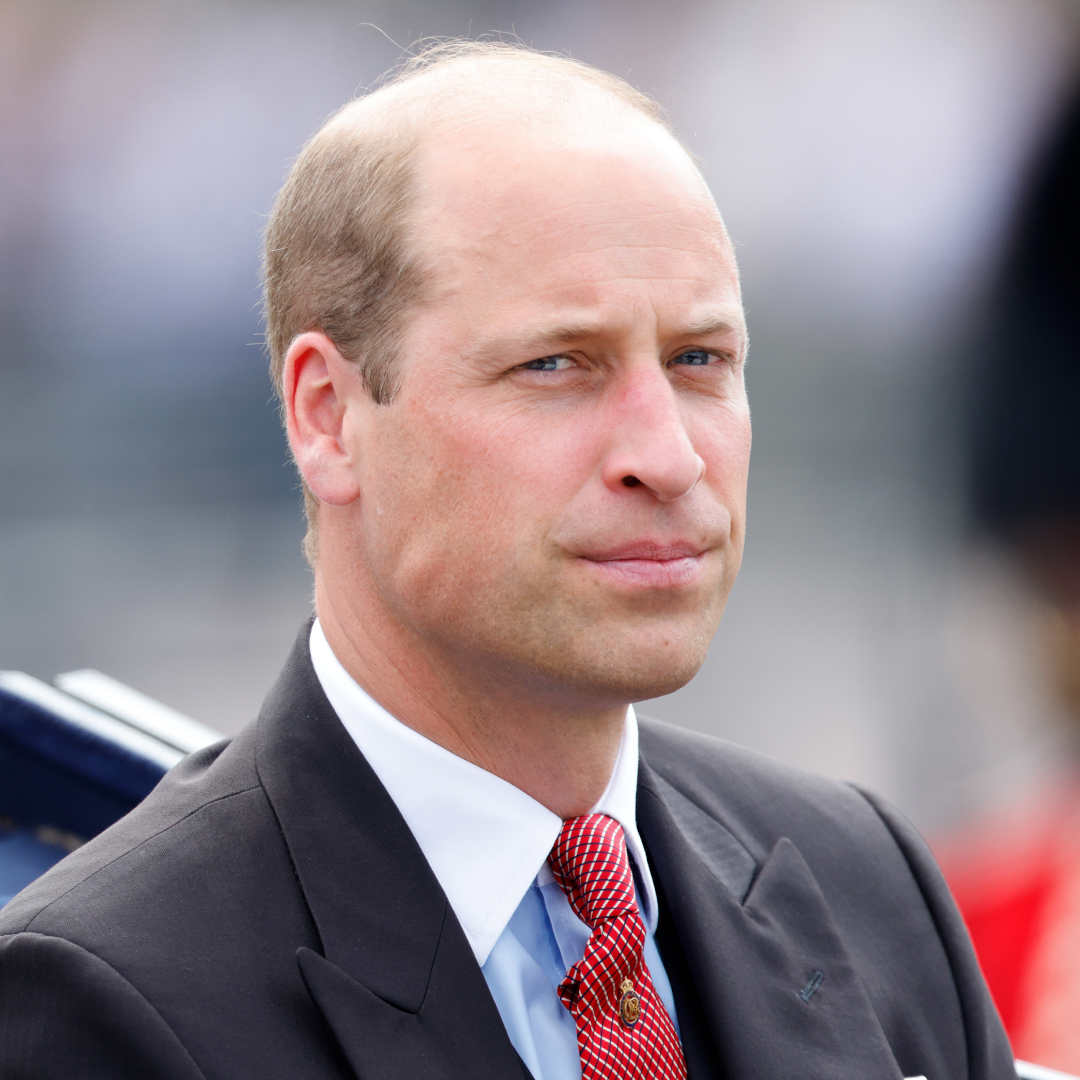 Prince William wears a black suit with a red tie and looks serious