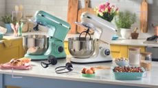 Yellow kitchen with white marble counters showing Aldi stand mixers in green and cream on a kitchen island with baked goods in turquoise bowls and pink plates