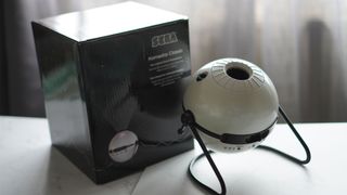 A white, spherical projector is placed on black legs on a white surface next to a black product box with white curtains behind it.
