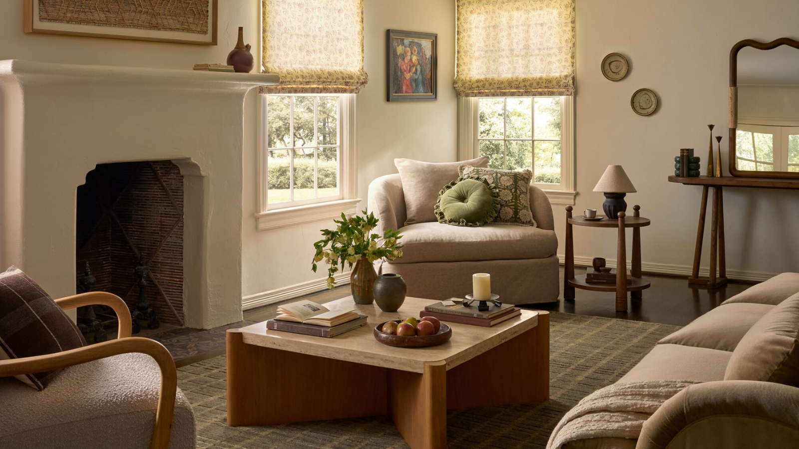 a living room with a large fireplace, a square coffee table, an accent chair with pillows on it, a small side table, and fabric blinds in front of the windows in the corner
