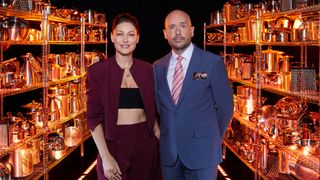 Cooking With The Stars 2025 Hosts Emma Willis and Tom Allen standing side by side, superimposed on a black background with rows of pots and pans behind them.