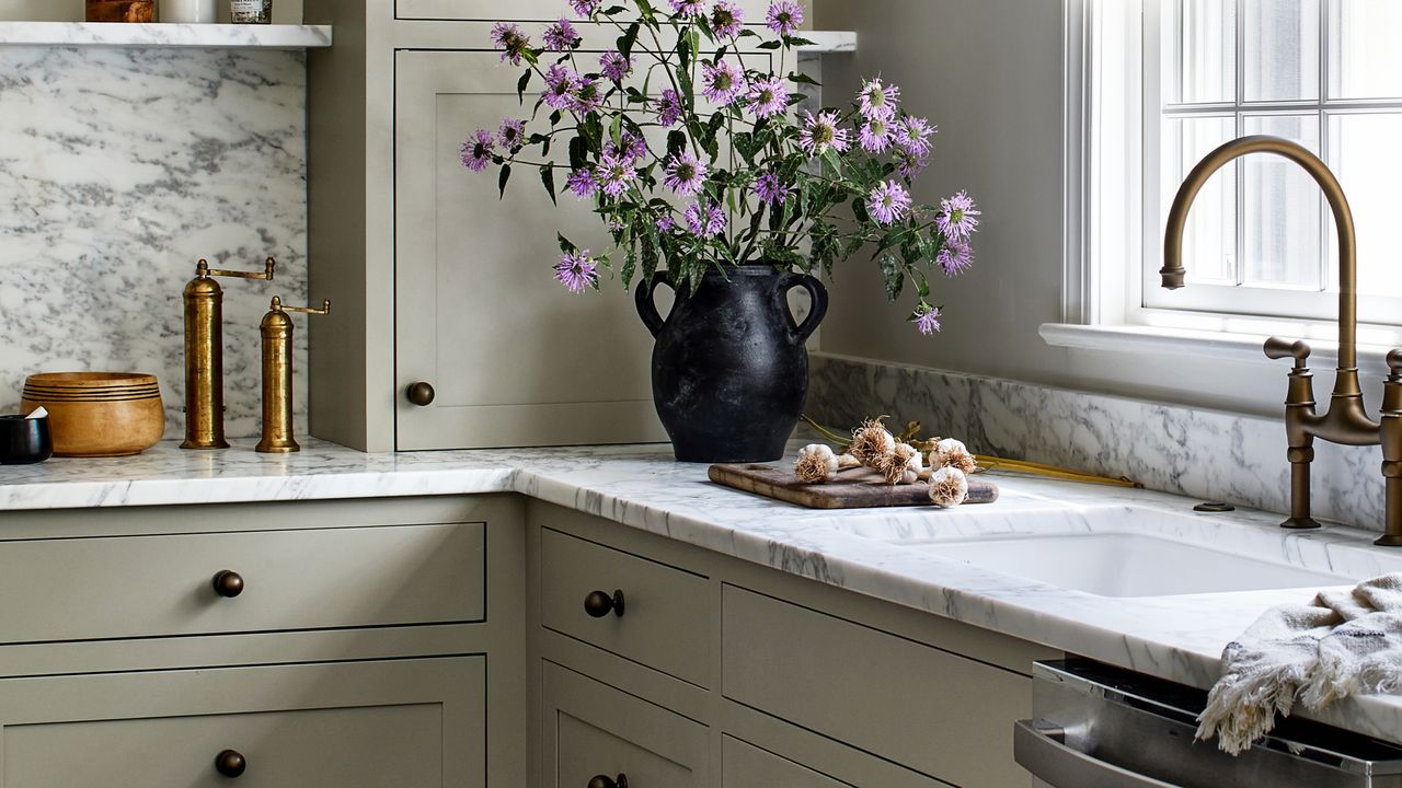 Close-up image of a kitchen counter top that is white and gray marble with light sage green cabinetry. There is a black vase with purple flowers in it and a cutting board with garlic on it. 