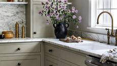 Close-up image of a kitchen counter top that is white and gray marble with light sage green cabinetry. There is a black vase with purple flowers in it and a cutting board with garlic on it.