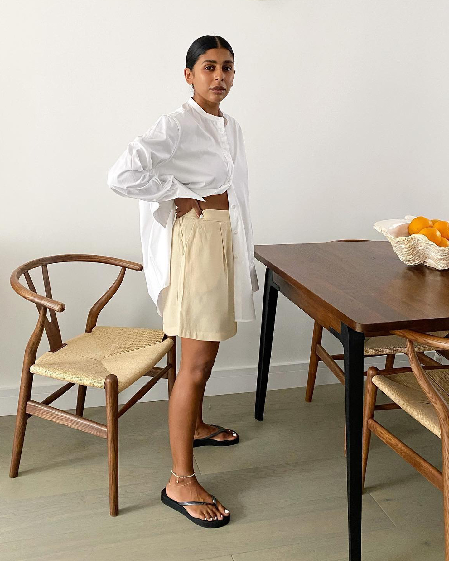 British style influencer Monikh Dale poses in her chic London dining room with an abstract white bowl full of oranges on the table wearing a white oversize line button-down shirt, butter-yellow shorts, anklets and black flat flip-flop sandals