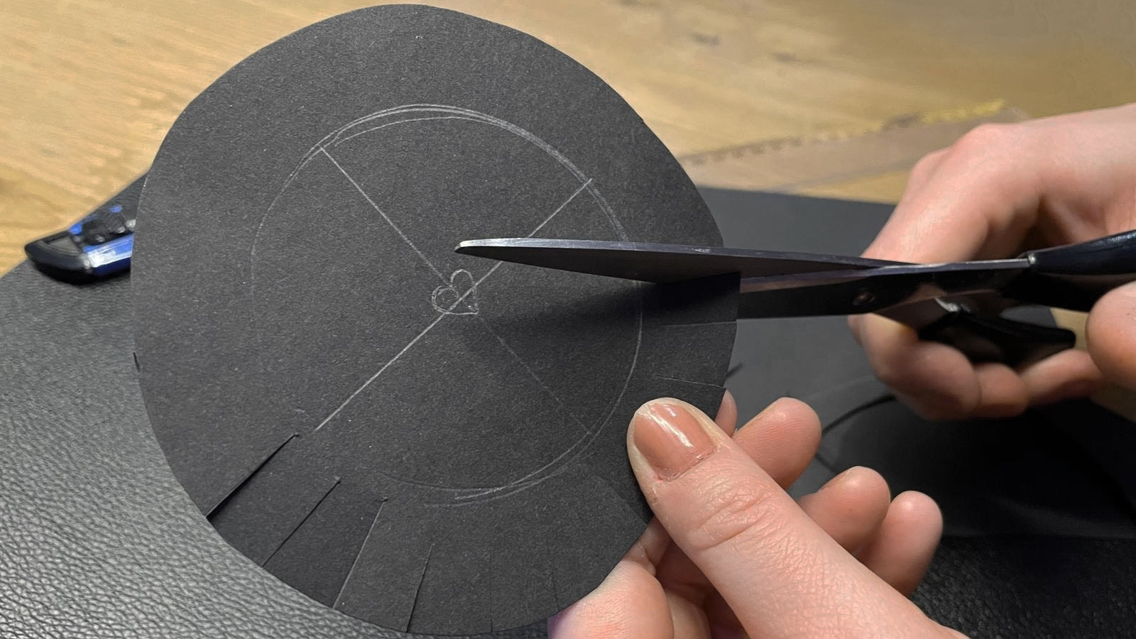 A person cuts a black circular piece of cardstock with scissors, revealing a heart shape in the center, on a wooden table