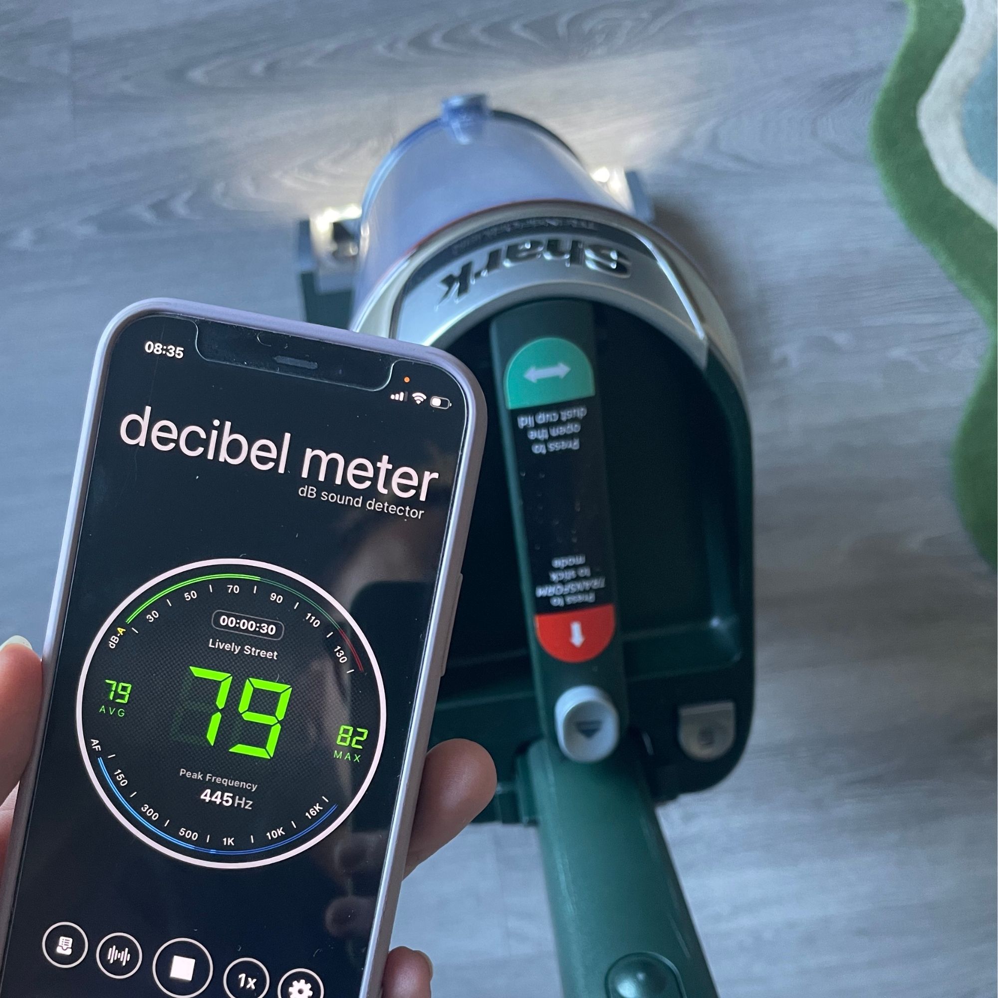 Shark PowerDetect Transformer Corded Upright Vacuum Cleaner on grey flooring, next to a hand holding up a decibel meter app, with 79 decibels showing on the screen