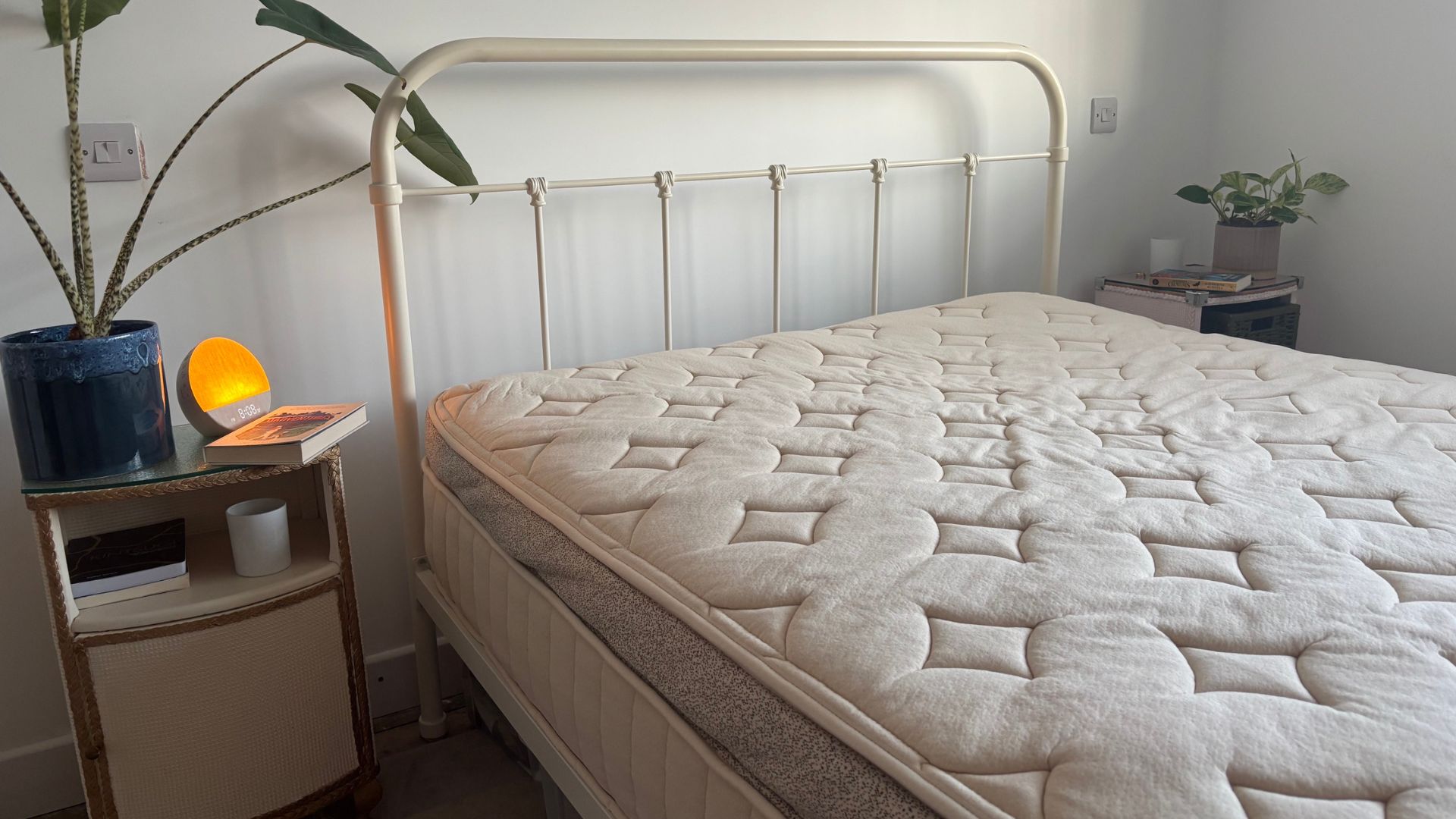 The image shows a beige mattress on a white bed frame, placed inside a neutrally decorated bedroom