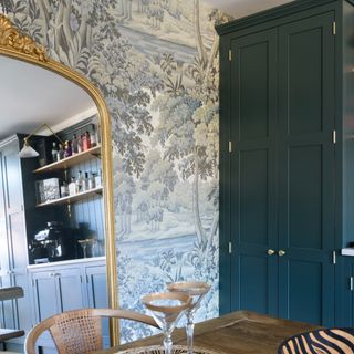 A dining room with a large leaning mirror and a vintage-style nature-inspired wallpaper mural