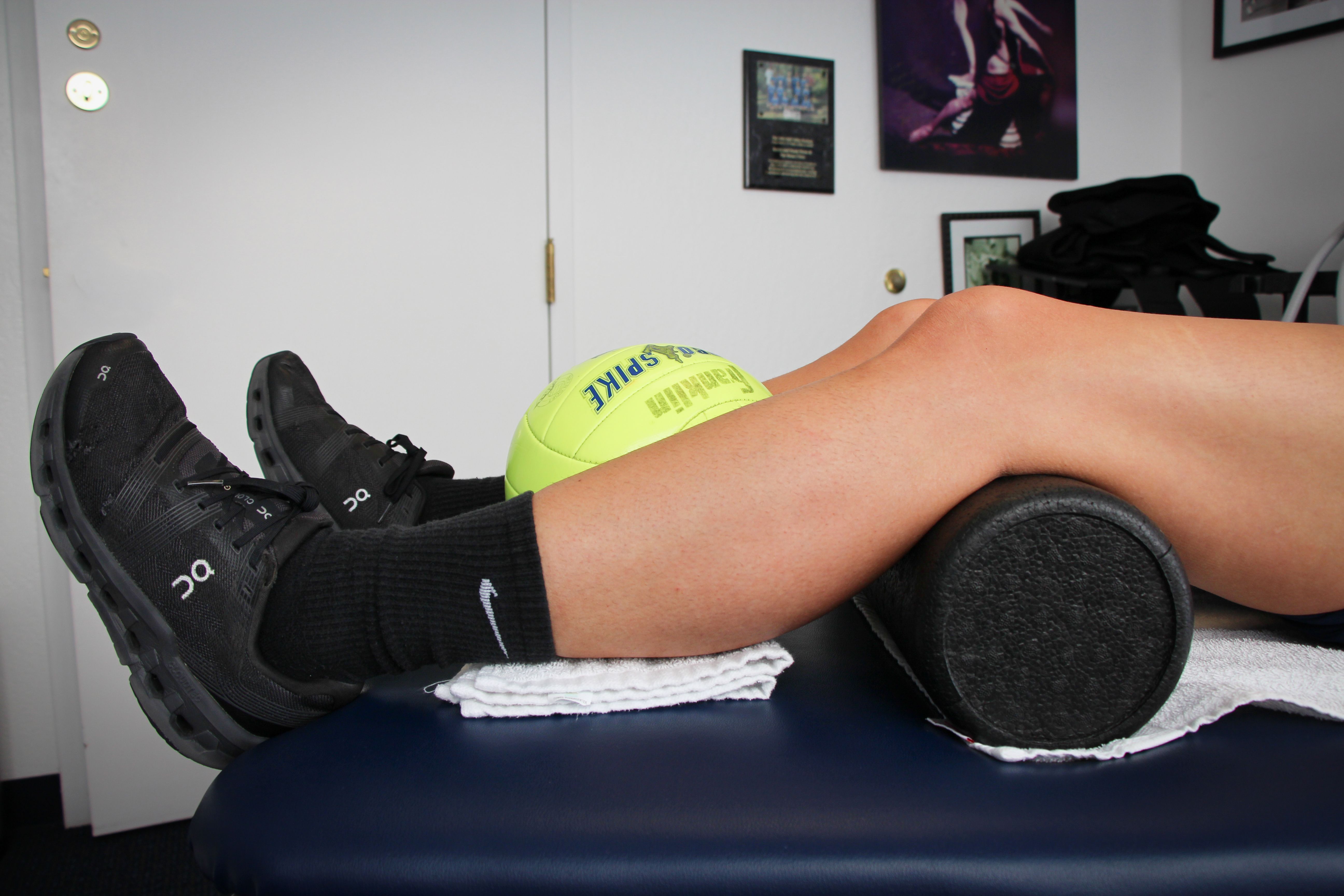Image of legs on a treatment table, a foam roller under the knees and a yellow ball held between the lower legs. Heels are resting on the table. 