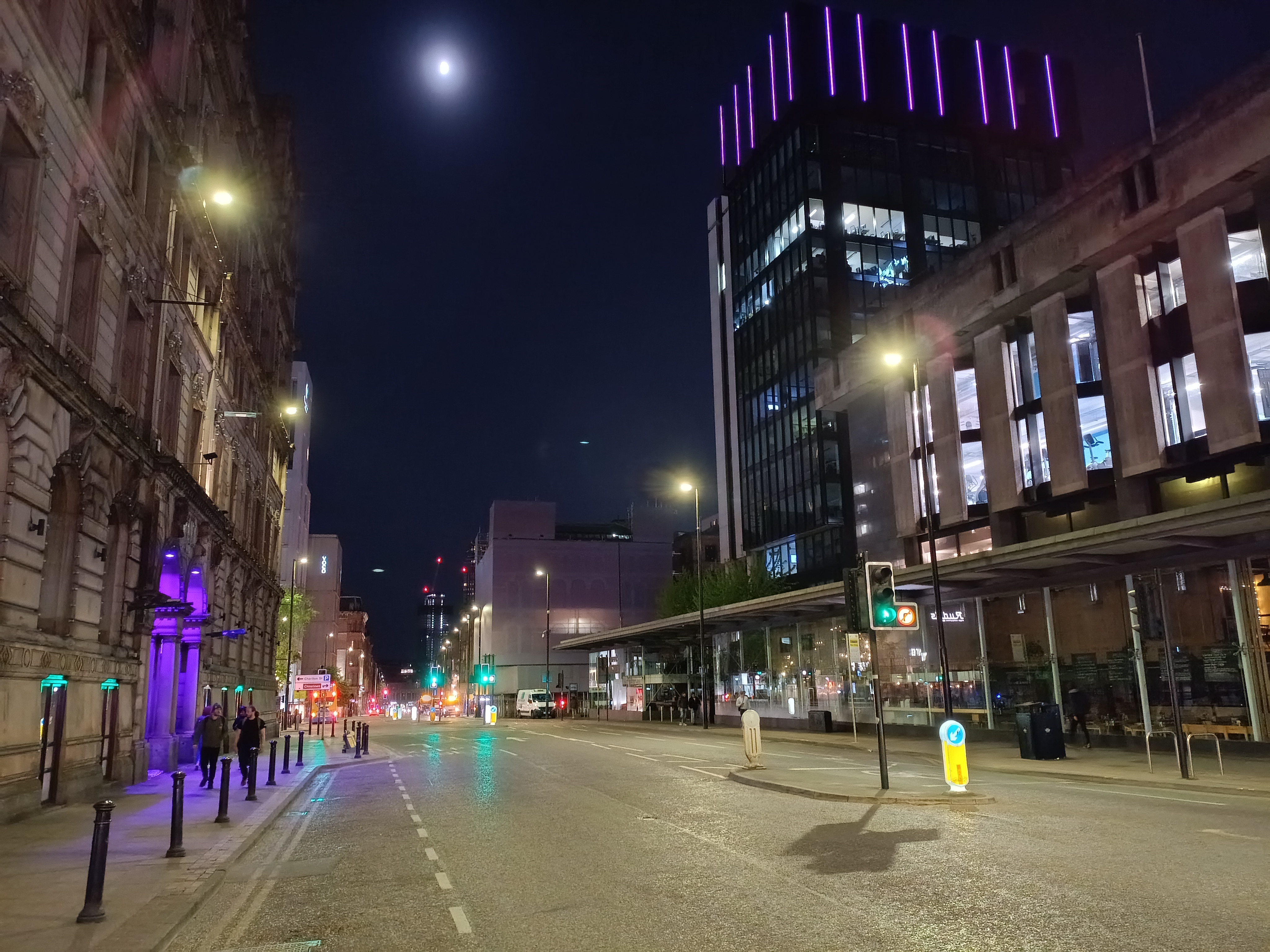 A quiet city street at night with a full moon visible in the deep dark sky above tall buildings.