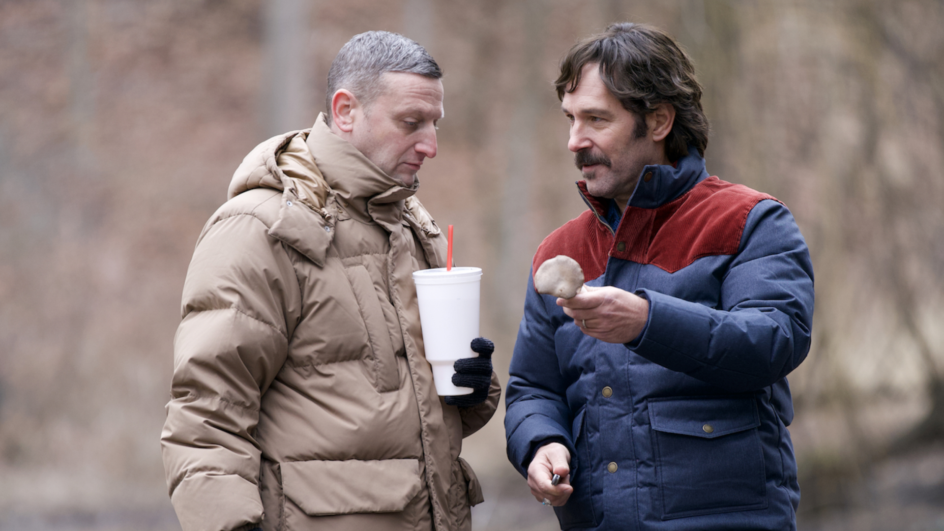 tim robinson wears a brown coat and holds a big gulp cup while looking at a mushroom with paul rudd in a blue and red coat in the movie friendship