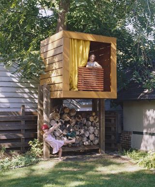 Children play at treehouse with wooden slat design, and log store below