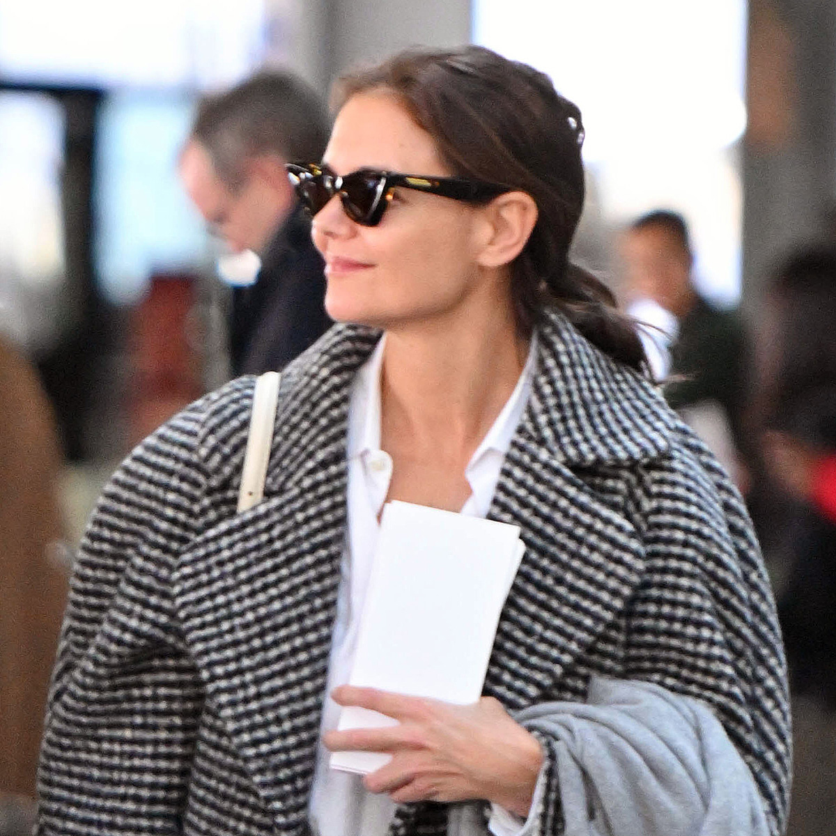Katie Holmes at JFK airport wearing a printed black-and-white checked coat over a white button-down shirt with wide-leg jeans and black loafers