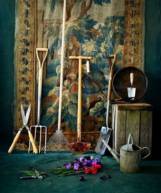 A tapestry wall hanging with an assortment of garden tools and a watering can leaning on it.