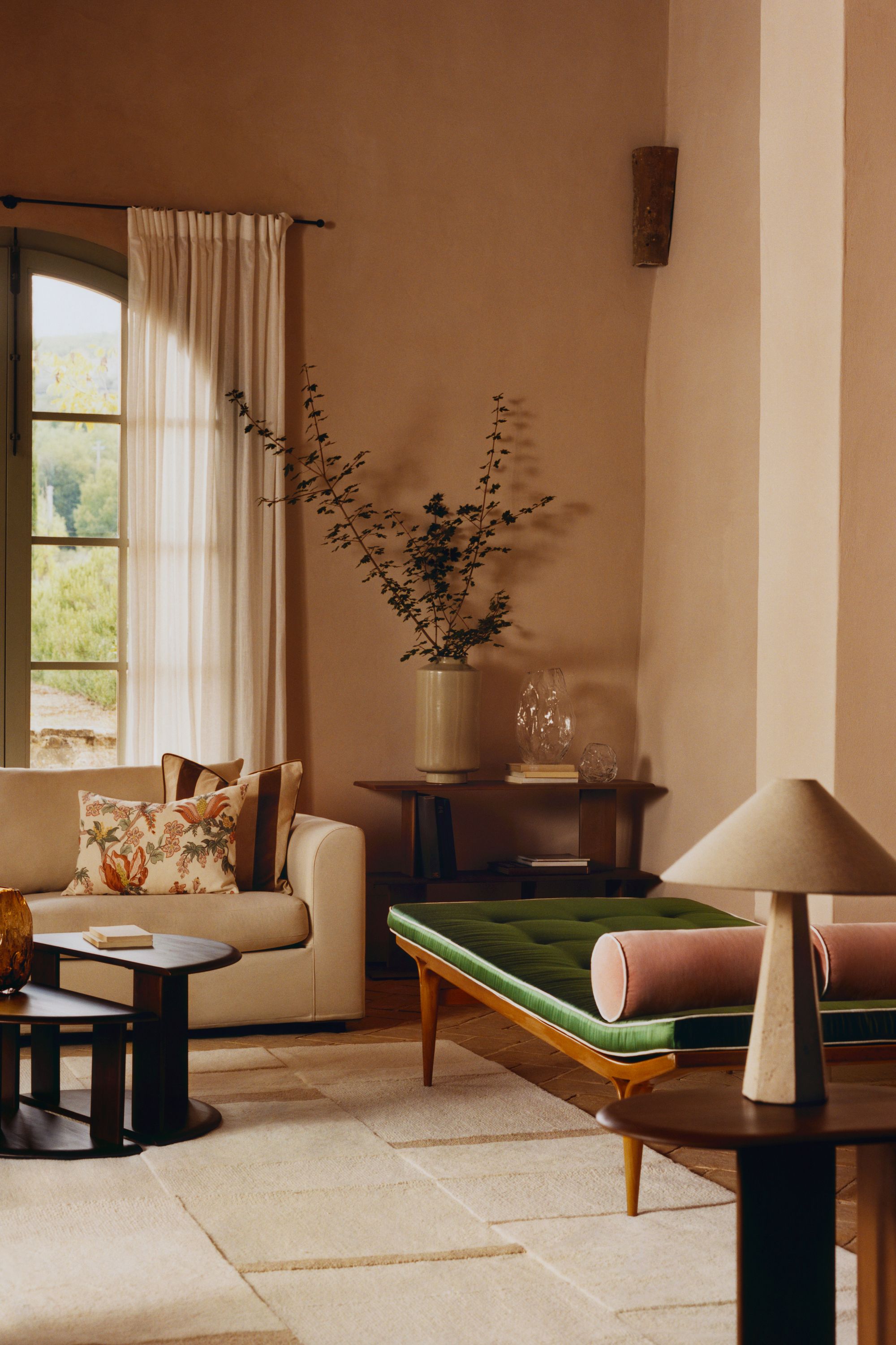 Image of a beige living room with an off-white rug, off-white sofa, dark wood nesting coffee table, and a green velvet and wood daybed. There is a window at the back with a sheer curtain.