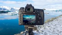 Camera on tripod capturing a snowy icy landscape with a calm blue lake and mountains in the background on a clear day