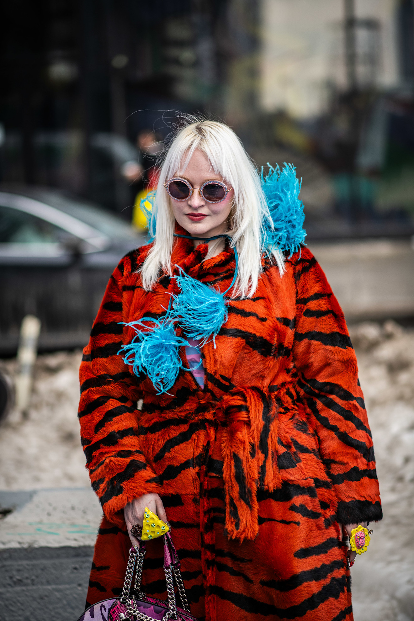 a woman with platinum blonde hair with sunglasses and a red and black coat with blue feathers