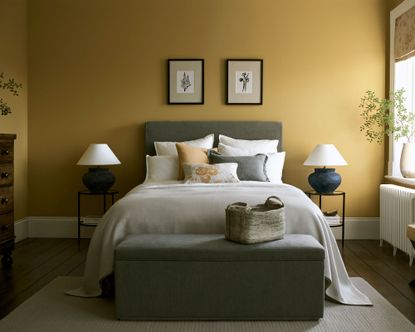 Saffron yellow painted bedroom, dark wooden flooring, gray upholstered bed, white linen with gray and yellow cushions, matching bedside lamps on metal and glass tables, two floral prints mounted on wall above bed