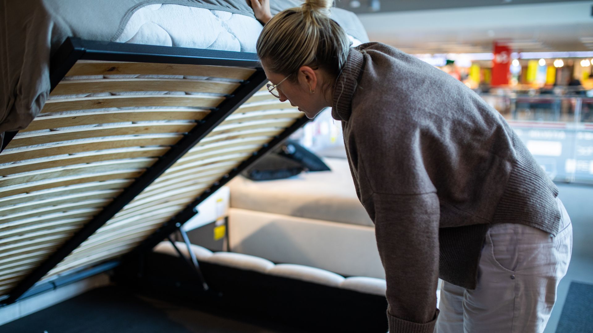 A blonde woman lifts up the storage compartment of a storage bed in a furniture store.