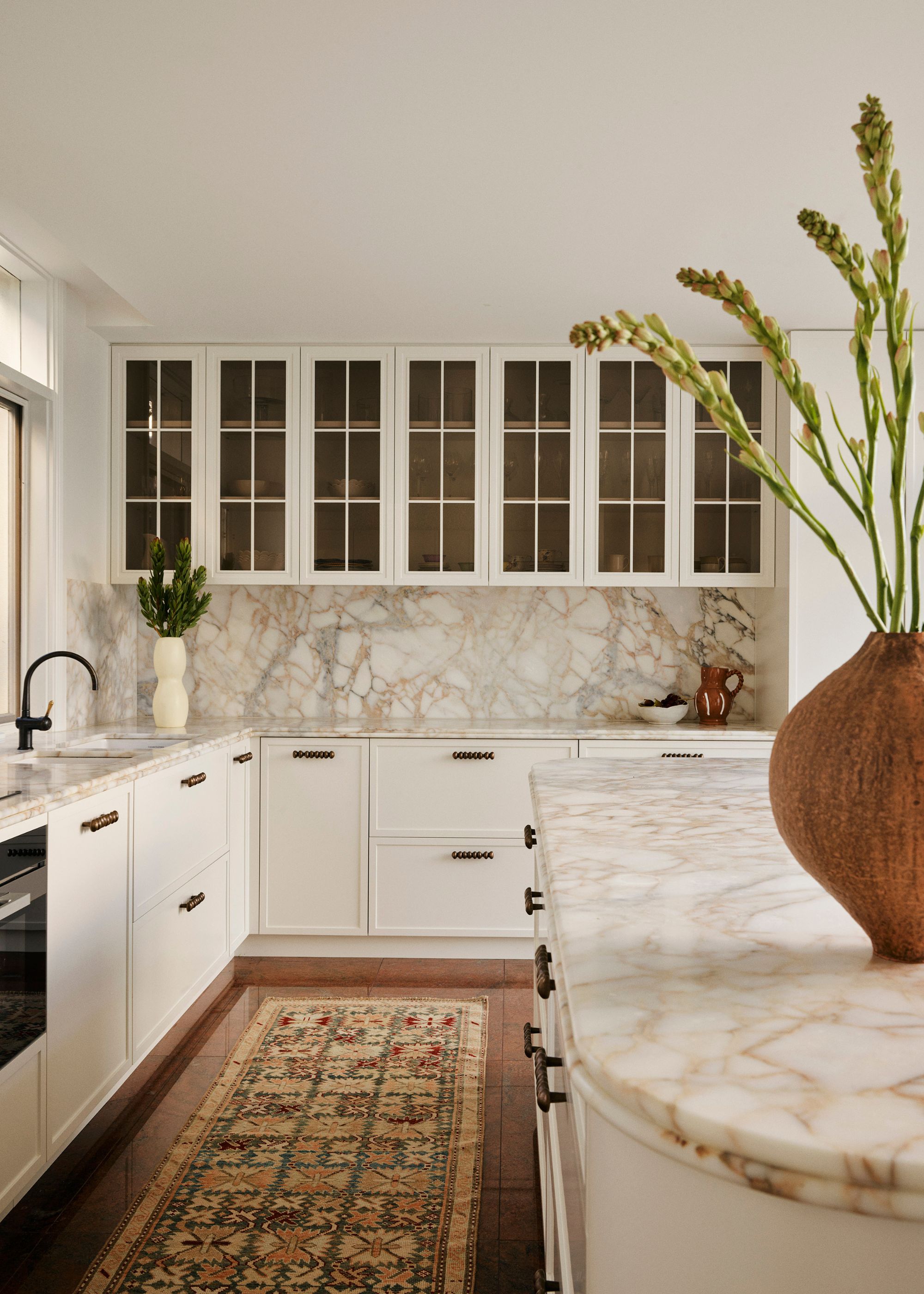 A white kitchen, a stone backsplash, a patterned runner, a vase of stems, and brass hardware