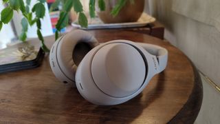 A white pair of Sony WH-1000XM4 headphones on a wooden table with a plant in the background.