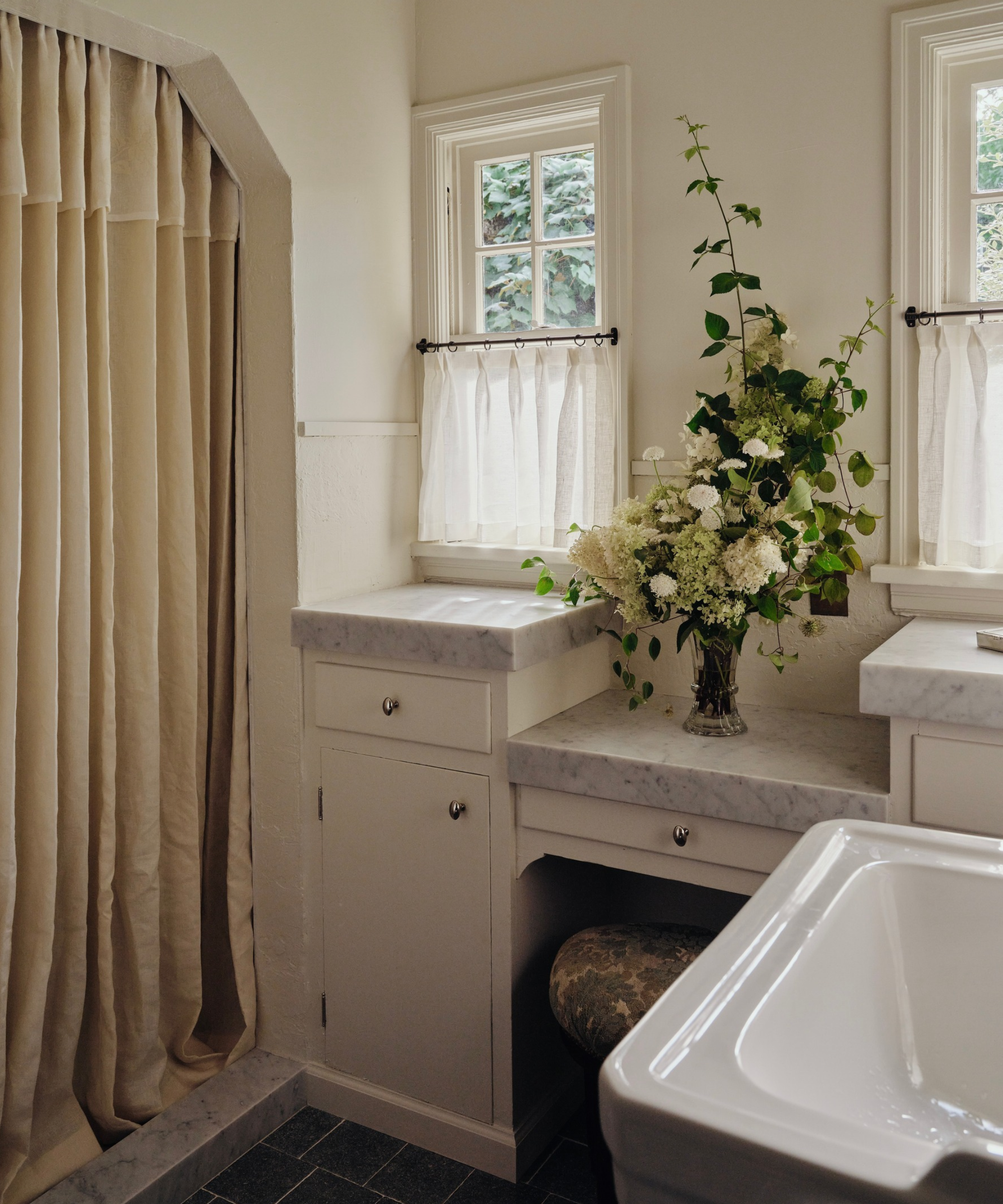 Light, bright bathroom featuring gray marble countertops, creamy sheer cafe curtains, and an overflowing vase of wild flowers.