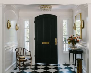 An entryway with black and white chequerboard floor, white walls, black door, gold mirror and black console table