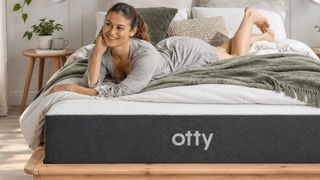 A dark haired woman lies on the charcoal and white Otty Original Ice Hybrid Mattress, which is placed on a wooden bed frame and set within a neutrally decorated bedroom