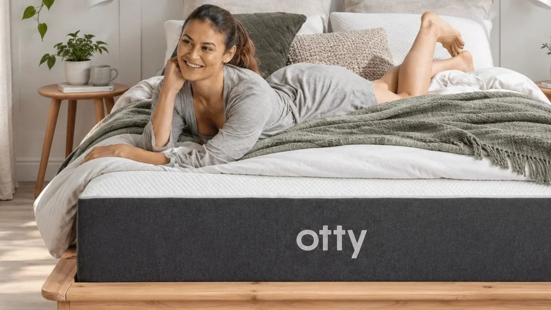 A dark haired woman lies on the charcoal and white Otty Original Ice Hybrid Mattress, which is placed on a wooden bed frame and set within a neutrally decorated bedroom