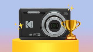 A Kodak Pixpro FZ55 compact camera with a zoom lens displayed on a pedestal, surrounded by stars and a trophy, set against a soft pastel background