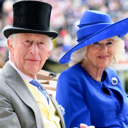 King Charles wears a gray suit with a black top hat, while Queen Camilla wears a large royal blue hat with a matching jacket