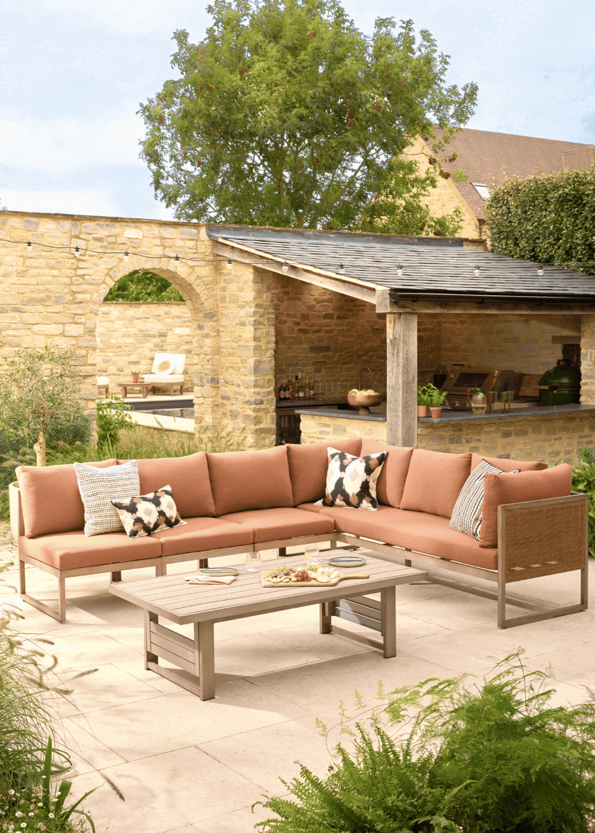 A gif showing three versions of Next's modular outdoor sofa and table lounge set in rust in a modern patio with greenery all around, a brick wall with an arch leading to another part of the garden and a covered outdoor kitchen in the back corner