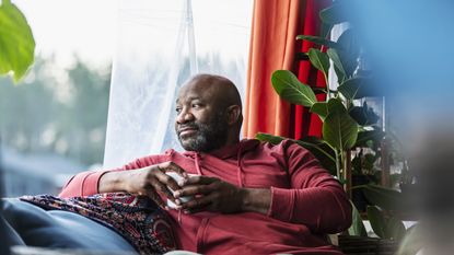 An older man with a cup of coffee sits on his sofa and looks out the window, looking relaxed.